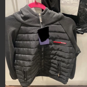 Svart quiltad jacka med huva Paris - Svart jacka med quiltad front, huva och dragkedja hela vägen. Jackan har två fickor med dragkedja och en diskret röd Paris-logga på bröstet. Ärmarna är släta och materialet känns mjukt och sportigt. Perfekt för kyliga dagar.