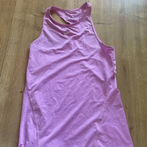 Nike rosa Dri-Fit träningsklänning tennis - Nike Dri-Fit klänning i en snygg ljusrosa färg, perfekt för träning. Modellen är kort och ärmlös med racerback och har ett sportigt, tight passform. Materialet är lätt och andas, tillverkat i polyester för maximal komfort under aktivitet.