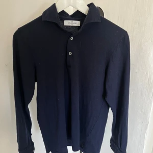 Mörkblå pikétröja från Gran Sasso - Passar S men är liten i storlekn, Klassisk mörkblå pikétröja från Gran Sasso med lång ärm och krage. Tröjan har knäppning med två knappar framtill och muddar med knapp vid ärmslut. Tillverkad i mjuk ull som ger en skön känsla och stilren look. Hör av dig om du har frågor!🙌