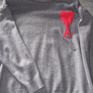 Grå stickad tröja från Ami Paris - Säljer en grå stickad tröja från Ami Paris med deras ikoniska röda hjärta och A broderat på bröstet. Tröjan har rund hals, långa ärmar och ribbade muddar. Perfekt för dig som gillar stilrena plagg med en twist.
