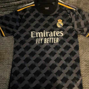 Real Madrid svart matchtröja Adidas - Snygg svart Real Madrid fotbollströja från Adidas med grått grafiskt mönster och gula detaljer på axlarna. Tröjan har klubbmärket broderat på bröstet och Emirates Fly Better-tryck framtill. Tillverkad i lätt och ventilerande polyester, perfekt för träning eller match.