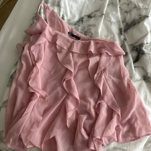 Rosa volangtop från Zara oneshoulder - Supersöt rosa topp från Zara i oneshoulder-modell med tunna axelband och volangdetaljer framtill. Luftigt tyg som ger en flowy känsla, perfekt för varma dagar. Toppen är ärmlös och har en feminin look med volanger längs hela framsidan.