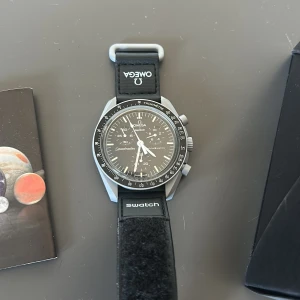 Omega x Swatch MoonSwatch Speedmaster - Omega x Swatch Speedmaster MoonSwatch 'Mission to the Moon' med svart urtavla, grå boett och svart textilrem. Klockan har kronograf, tachymeter och coola detaljer som månmotiv på baksidan. Levereras i originalförpackning.
