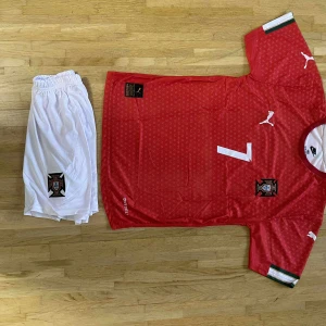 Portugal Ronaldo fotbollströja Puma - Portugal fotbollströja med Ronaldo och nummer 7, röd med små vita mönster och vita detaljer. Puma-logga på bröst och ärm, landslagsemblem på bröstet. Kortärmad modell i lätt funktionsmaterial. Matchande vita shorts med emblem ingår.