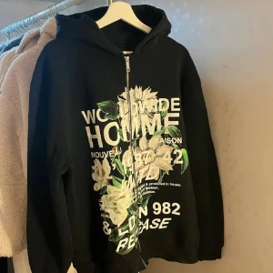 Svart hoodie med blommor från BoohooMAN - Svart hoodie från BoohooMAN med dragkedja, huva och stora vita blommor samt texttryck på framsidan. Snygg streetstil med gröna och vita detaljer. Perfekt för dig som vill sticka ut med en cool och unik look.