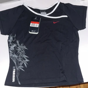 Svart träningstopp från Nike, storlek 152-158 - Nike träningstopp i svart med vit kant vid halsen och ett rött Nike Swoosh på bröstet. Toppen har korta ärmar och ett stilrent tryck med grå växtmotiv på sidan samt texten 'Training'. Skön passform och sportig look, perfekt för gymmet.
