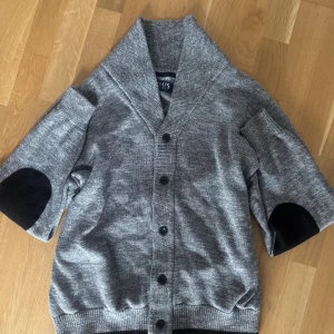 Grå kofta från Chaps med knappar - Snygg gråmelerad kofta från Chaps med bred krage och svarta knappar framtill. Koftan har trekvartsärmar med svarta armbågslappar och ribbade muddar. Perfekt för lager-på-lager och chill stil.