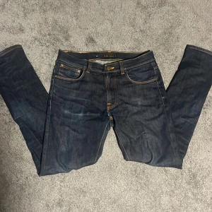 Mörkblå jeans från Nudie Jeans 32/34 - Säljer ett par mörkblå jeans från Nudie Jeans i storlek 32/34. Klassisk femficksmodell med orange kontrastsömmar och Nudie-logga på bakfickorna. Jeansen har rak passform och är tillverkade i slitstarkt denimtyg.