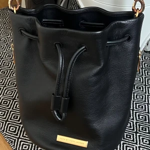 Svart bucket bag från Marc Jacobs - Snygg svart bucket bag från Marc Jacobs med guldfärgade detaljer och snörning upptill. Väskan är tillverkad i mjukt skinn och har en stilren, rundad form. Perfekt för dig som gillar minimalistisk design och vill ha plats för allt du behöver. Kan även tänka mig att byta mot annan väska ifrån Marc Jacobs 