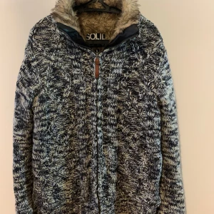 Stickad teddyjacka från Solid - Mysig stickad teddyjacka från Solid med grovt mönster i svart, grått och vitt. Jackan har fluffigt foder och huva i beige teddy, dragkedja med läderdetalj och ribbade muddar. Perfekt för kalla dagar och har en riktigt chill vibe.