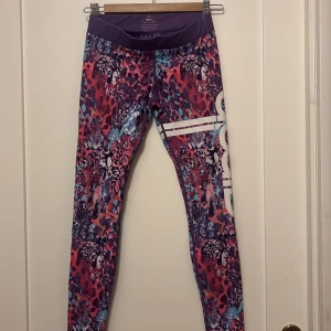 AIM'N tights XS - Färgglada tights från AIM'N. De har ett mönster i lila, rosa, blått och vitt med vita ränder och logga på benet. Låg midja och stretchigt material i polyester och spandex. Perfekta för träning och att sticka ut på gymmet.