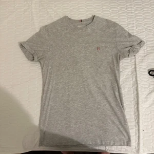 Grå t-shirt från Les Deux - Snygg grå t-shirt från Les Deux med diskret röd II-logga broderad på bröstet. Klassisk rund hals och korta ärmar. Tillverkad i mjuk bomull som känns skön mot huden. Perfekt för dig som gillar stilren och enkel design.