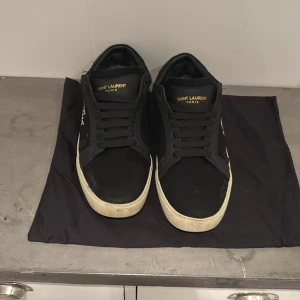 Svarta Saint Laurent sneakers - Svarta sneakers från Saint Laurent med vit logotyp på sidan och guldfärgad text på plösen. Skorna har snörning, rund tå och vit platt sula. Materialmix av skinn och textil ger en cool kontrast. Perfekt för dig som gillar stilrena och lyxiga street-vibes.