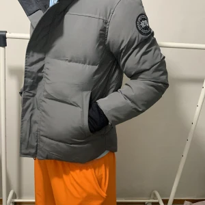 Grå pufferjacka från Canadian Goose - Grå pufferjacka från Canadian Goose med huva och svart patch på ärmen. Jackan har dragkedja och knappar framtill, två stora fickor och justerbar huva. Perfekt för kalla dagar och har en clean, modern look som funkar till streetwear.