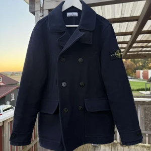 Mörkblå skepparkavaj från Stone Island - Stilren mörkblå skepparkavaj från Stone Island med klassisk dubbelknäppning, bred krage och två stora fickor framtill. Jackan har ikonisk logotyp på ärmen och är tillverkad i ull för en varm och exklusiv känsla. Nypris 18 000kr