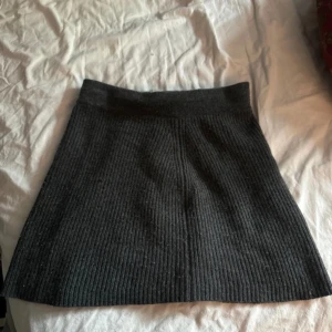 Mörkgrå ribbad stickad kortkjol - Säljer en mörkgrå stickad kortkjol med ribbad struktur. Kjolen har en enkel och stilren design med bred resår i midjan och lätt utsvängd form. Perfekt att matcha med en oversized tröja eller boots för en chill look.