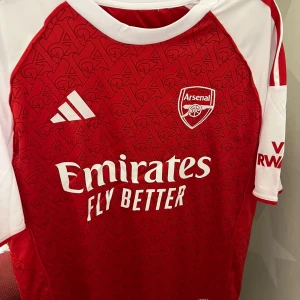 Arsenal hemmatröja Adidas röd/vit - Säljer en officiell Arsenal hemmatröja från Adidas. Tröjan är röd med vita ärmar och har ett diskret mönster med bokstaven A över hela framsidan och ryggen. Klassiska Adidas-ränder på axlarna, Arsenal-logga och sponsortryck framtill. Tillverkad i lätt och ventilerande material.