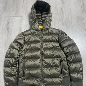 Olivgrön dunjacka Parajumpers Sheen - Snygg olivgrön dunjacka från Parajumpers, modell Sheen. Jackan har huva, ribbade muddar och dragkedja framtill. Klassisk PJS-logga på ärmen och två dragkedjeförsedda fickor. Perfekt för kalla dagar och riktigt street. Storlek Young large (passar även xs-s)