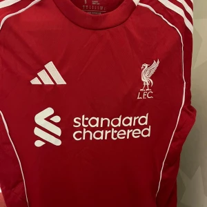 Liverpool FC röd matchtröja Adidas S - Liverpool FCs officiella matchtröja i rött från Adidas med vita detaljer och logga. Tröjan har korta ärmar, rund hals och är tillverkad i lätt, ventilerande funktionsmaterial. Tryck med Standard Chartered och klubbmärke på bröstet.