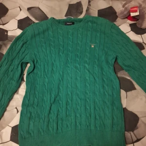 Grön kabelstickad tröja från Gant - Snygg grön kabelstickad tröja från Gant med rund halsringning och diskret logga på bröstet. Tröjan har långa ärmar och ribbade muddar vid ärmslut och nederkant. Perfekt för lager-på-lager och chill höst- och vinterstil.