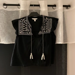Svart blus med broderi från H&M - Svart blus från H&M med vit broderad detalj runt halsen och axlarna. Blusen har korta volangärmar och snörning med tofsar framtill. Materialet är mjukt och lätt, perfekt för en chill look. Snygg kontrast mellan svart tyg och vita broderier.