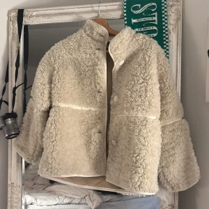 Beige teddyjacka från H&M - Supermjuk och fluffig teddyjacka från H&M i beige färg. Jackan har hög krage, stora knappar framtill och är helfodrad. Perfekt för kalla dagar och ger en riktigt cozy vibe. Passar dig om du är storlek 140