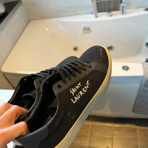 Svarta Saint Laurent sneakers - Snygga svarta sneakers från Saint Laurent med vit handskriven logga på sidan. Skorna har klassisk låg profil, svarta snören och vit platt sula. Ovandelen är i canvas och detaljer i skinn, vilket ger en stilren och trendig look.