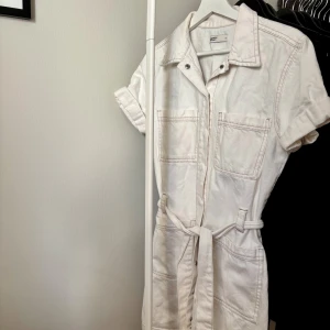 Vit jeansklänning från Ginatricot  - Vit kort jeansklänning från Ginatricot med korta ärmar, krage, två stora bröstfickor och knytbälte i midjan. Klänningen har dragkedja framtill och markerade sömmar för en cool look. Perfekt för dig som gillar stilrena och avslappnade outfits. En stor favorit som tyvärr är för liten:(