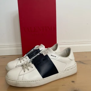 Valentino Garavani sneakers vit/blå - Säljer ett par snygga Valentino Garavani sneakers i vitt skinn med bred marinblå detalj över mitten. Klassisk låg modell med snörning och coola nitar på hälen. Kommer med originalröd box. Perfekta för dig som vill ha något exklusivt och stilrent.
