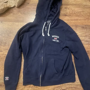 Morris hoodie  - Mysig marinblå hoodie från Morris med vit dragsko i huvan och broderad logga på bröstet. Tröjan har hel dragkedja, kängurufickor och mjukt bomullsmaterial. Perfekt för chill dagar eller när du vill ha en klassisk streetstyle-look.