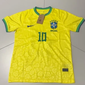 Brasilien Neymar Jr #10 fotbollströja Nike - Gul Brasilien fotbollströja från Nike med gröna och blå detaljer, nummer 10 och Neymar Jr på ryggen. CBF-emblem på bröstet, korta ärmar och rund hals med knapp. Tillverkad i lätt Dri-FIT material med subtilt mönster över hela tröjan.