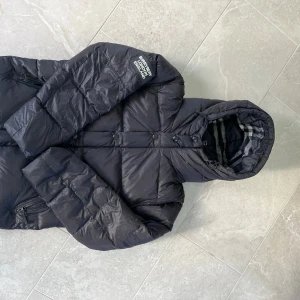 Svart Burberry pufferjacka med huva - Svart pufferjacka från Burberry London England med avtagbara ärmar, så du kan bära den som väst. Jackan har quiltad design, dragkedjor på fickorna, tryckknappar och en stor huva med rutigt foder. Perfekt för kalla dagar och riktigt snygg streetstil.