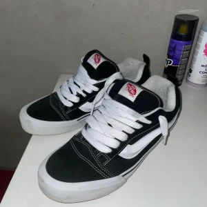 Svarta och vita Vans sneakers - Säljer ett par klassiska Vans sneakers i svart och vitt med den ikoniska sidostripen. Skorna har vit snörning, rund tå och platt sula med gummisula i brunt. Materialet är en mix av canvas och mocka, och de har en cool streetstil med 'Off The Wall'-logga på tungan.