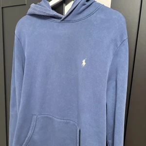 Blå hoodie från Polo Ralph Lauren - Snygg blå hoodie från Polo Ralph Lauren med klassisk broderad logga på bröstet. Tröjan har huva, känguruficka och är tillverkad i mjuk bomull. Okej skick då den är använd ganska mycket och ganska urtvättad 
