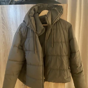 Olivgrön pufferjacka från Zara - Säljer en olivgrön pufferjacka från Zara med huva och snörning. Jackan har quiltad design, dragkedja framtill och långärmade ärmar. Perfekt för kalla dagar och har en loose passform som ger en chill vibe.