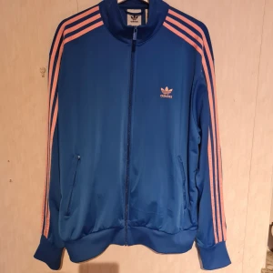 Blå Adidas track jacket med rosa ränder XL - Snygg blå Adidas track jacket med klassiska rosa ränder längs ärmarna och Adidas-logga på bröstet. Tröjan är endast testad så den är helt ny och otvättade! Strl: XL