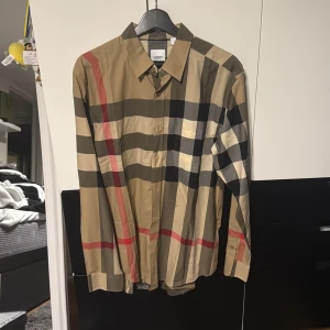 Rutig Burberry skjorta i beige och svart - Säljer en klassisk Burberry skjorta i storlek L med det ikoniska rutiga mönstret i beige, svart, röd och grå. Skjortan har lång ärm, knappar framtill och är tillverkad i mjuk bomull. Perfekt för dig som vill ha en stilren och tidlös look.