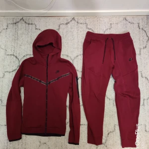 Mörkröd tracksuit från Nike - Snygga mörkröd tracksuit från Nike med svarta detaljer och snörning i midjan. Byxorna har en tapered passform, ribbade muddar vid bensluten och praktiska fickor. Perfekta för chill eller träning, tillverkade i mjukt bomullsmaterial. Byxorna är M & koftan är S. 