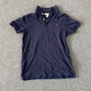 Marinblå pikétröja från L.O.G.G. - Snygg marinblå pikétröja från L.O.G.G. med klassisk krage och två knappar framtill. Tröjan har korta ärmar och en liten röd broderad detalj på bröstet. Tillverkad i mjuk bomull som är skön att bära hela dagen.