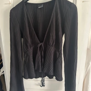 Svart ribbad omlottopp Gina Tricot - Svart långärmad omlottopp från Gina Tricot med ribbad struktur och knyt framtill. Toppen har djup v-ringning och är tillverkad i ett stretchigt material som sitter snyggt på kroppen. 