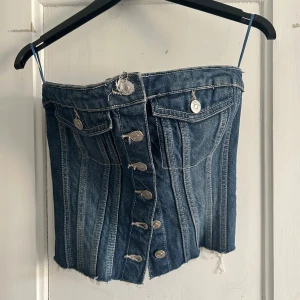 Jeans-korsett från Zara med knappar - Säljer en snygg jeans-korsett från Zara i blå denim med silverfärgade knappar framtill, råa kanter nertill och smockad baksida för extra passform. Toppen är axelbandslös och har dekorativa fickor framtill. 