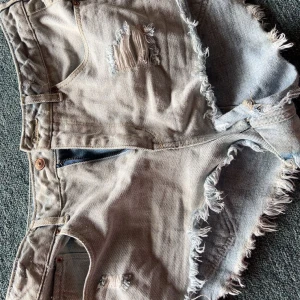 Rippade jeansshorts från H&M Divided - Säljer ett par ljusblå jeansshorts från H&M Divided med råa, fransiga kanter och slitna detaljer framtill. Klassisk femficksmodell med knapp och dragkedja. Perfekta för sommaren och ger en avslappnad vibe.