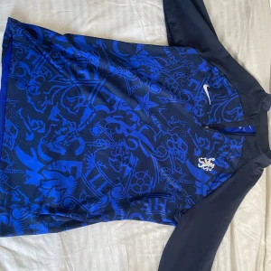 Blå långärmad Chelsea Nike Dri-Fit - Chelsea fotbollströja från Nike i slim fit-modell med lång ärm. Tröjan är blå med mörkblå detaljer och har ett unikt grafiskt mönster över hela framsidan. Tillverkad i Dri-Fit-material som andas och håller dig torr. Swoosh-logga och Chelsea-lejonet på bröstet.