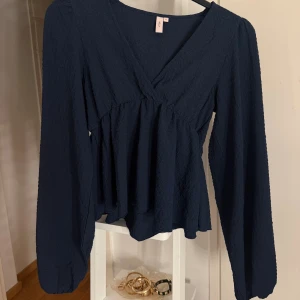 Mörkblå blus med v-ringning från Nelly - Supersnygg mörkblå blus från Nelly med v-ringning och långa ärmar. Blusen har en lätt strukturerad yta, markerad midja och volangig nederdel. Ärmarna efterliknar adoore klänningarnas ärmar!
