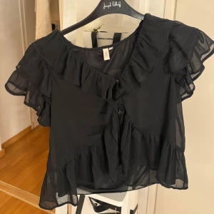 Svart volangblus från Nelly - Supersöt svart blus från Nelly med volanger längs ärmar, halsringning och nederkant. Blusen är kortärmad och har knappar framtill. Tillverkad i ett lätt och luftigt material. Den är använd fåtalgånger.