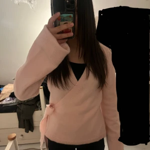 Ljusrosa omlottkofta med knyt - Säljer en superfin ljusrosa omlottkofta med breda långa ärmar och knyt i sidan. Koftan är stickad och har en v-ringad hals, perfekt för dig som gillar mjuka och mysiga plagg med lite extra stil. Passar dig som vill ha en chill och söt look.