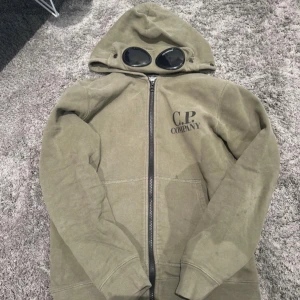 C.P. Company grön hoodie med goggles - Säljer en grön hoodie från C.P. Company med ikoniska goggles i huvan och dragkedja framtill. Tröjan har tryckt logga på bröstet. Ny pris 1700kr stl (152)