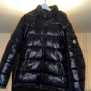 Svart Moncler pufferjacka med huva - Säljer en svart, glansig pufferjacka från Moncler med huva och dragkedja framtill. Jackan har två fickor med dragkedja och Moncler-logga på ärmen. Insidan har ett coolt serietryck. Perfekt för kalla dagar när du vill hålla stilen.