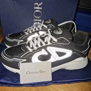 Svarta sneakers från Christian Dior - Snygga svarta sneakers från Christian Dior med vit kontrastdetalj och stora CD-loggor på sidorna. Skorna har snörning, rund tå och platt sula. Materialet är en mix av syntet och mesh för en sportig vibe. Perfekta för dig som gillar exklusiva märken och streetstyle.