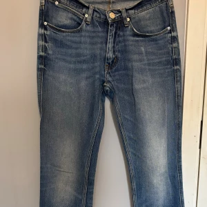  jeans från Acne Studios - Snygga blå jeans från Acne Studios med klassisk femficksdesign och raka ben. Jeansen har ljus tvätt med slitningar, silverfärgade nitar och knapp, samt brunt läderpatch bak i midjan. Perfekt för dig som gillar stilrena och tidlösa jeans.